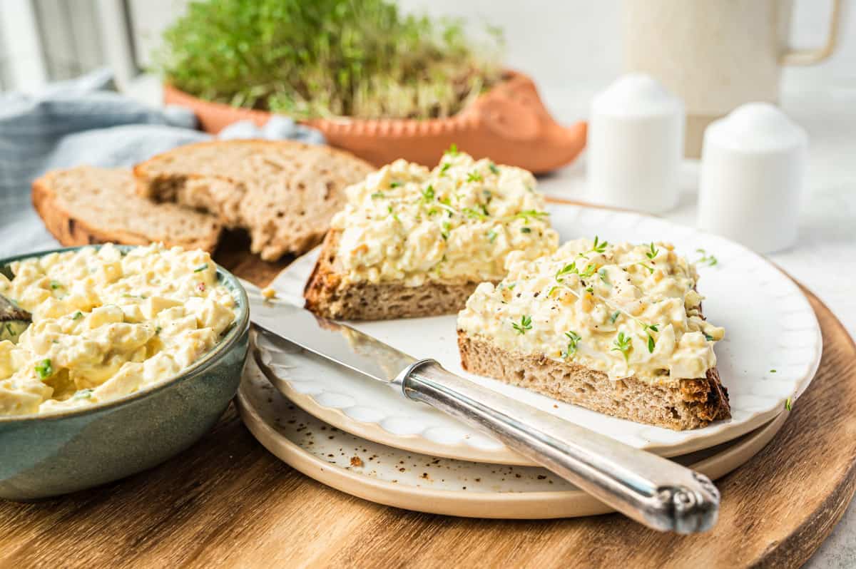 zwei halbe Scheiben Brot mit veganem Eiersalat auf weißem Teller mit Messer auf gedecktem Tisch