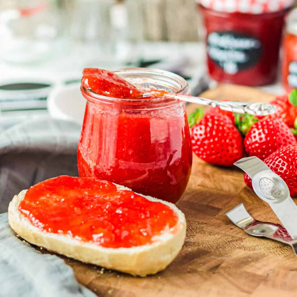 Ein halbes Brötchen mit Erdbeermarmelade, dahinter ein Marmeladenglas auf Holzbrett