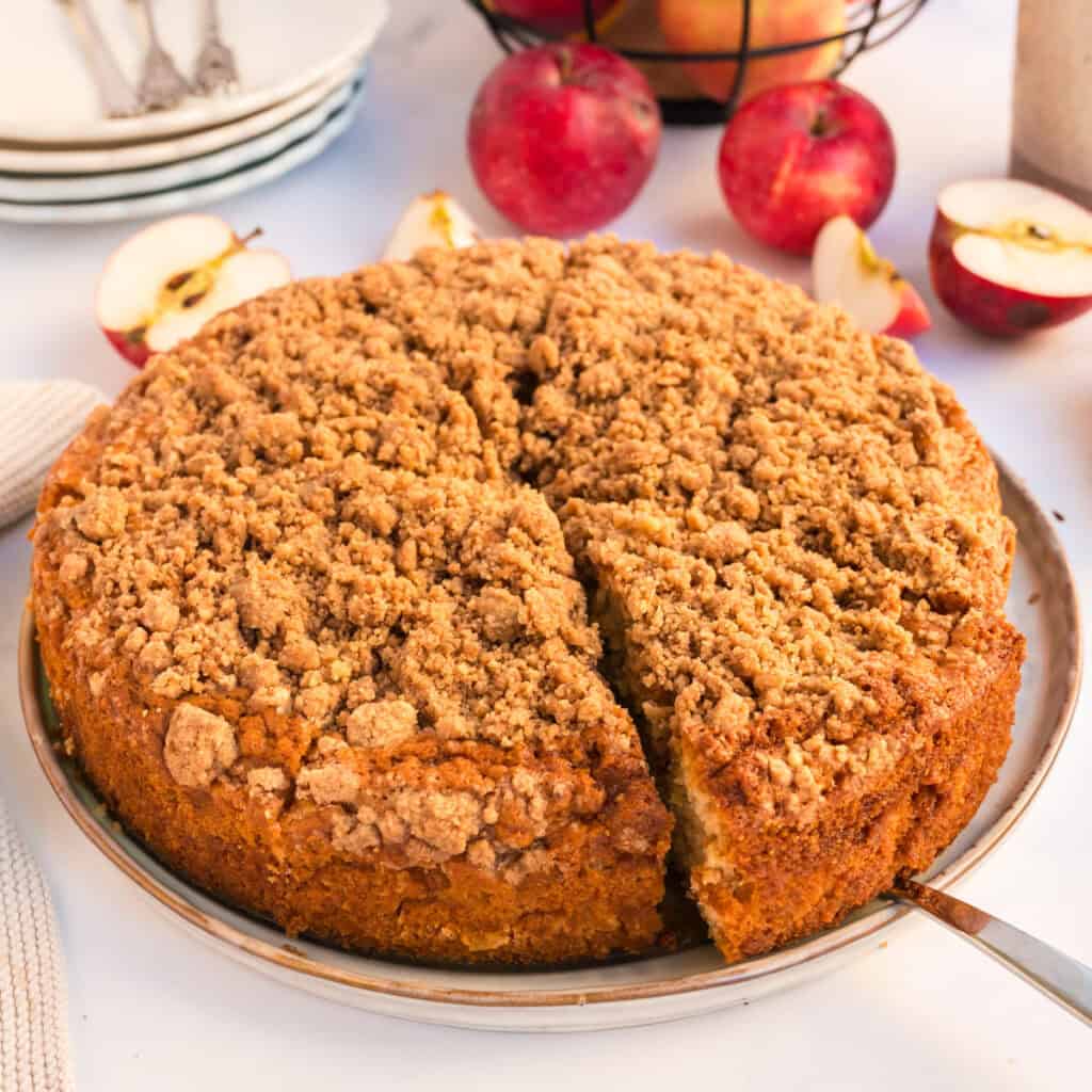 Angeschnittener ganzer Apfelkuchen mit Zimtstreuseln auf heller Platte, Kuchenheber unter einem Stück