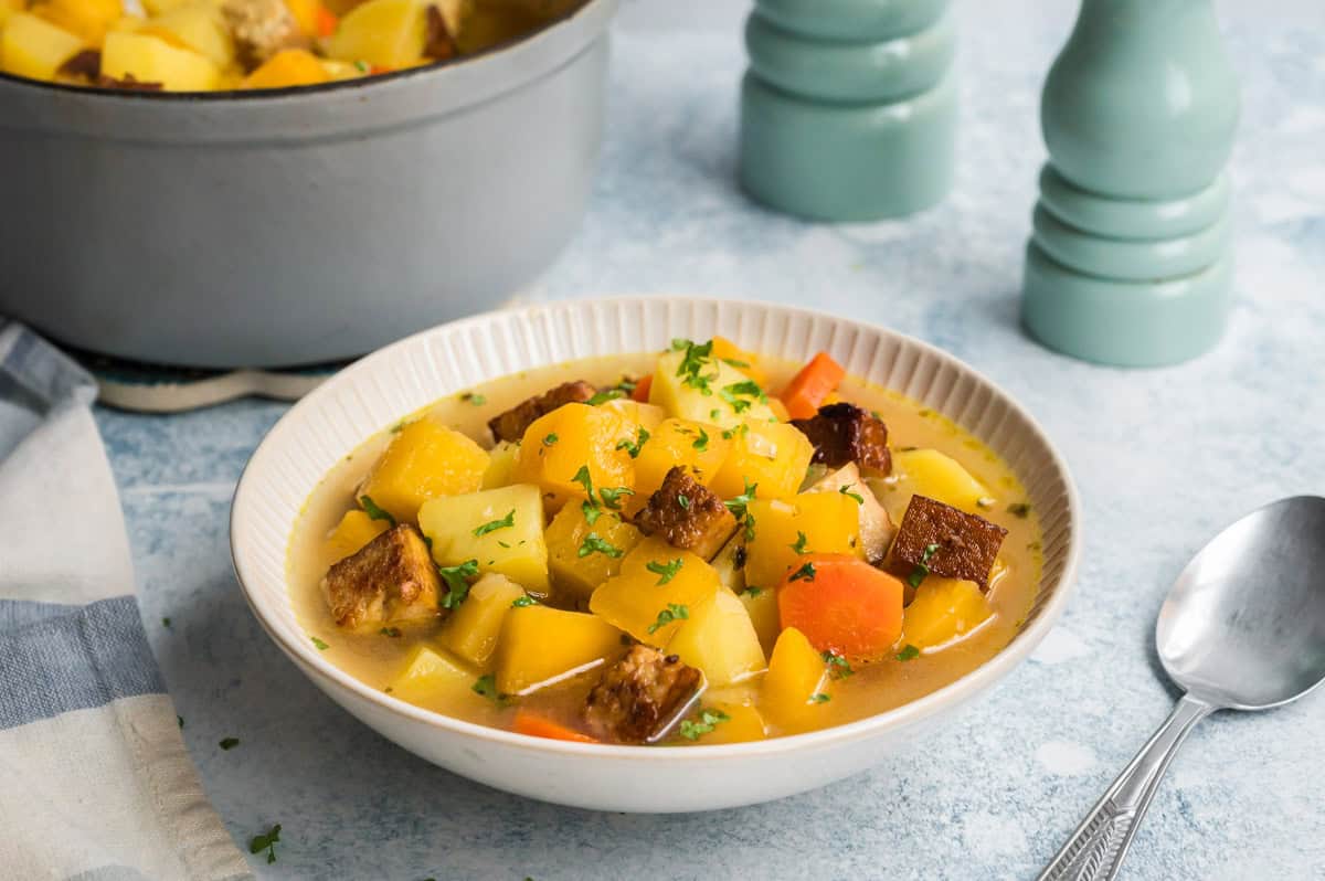 Steckrübeneintopf mit Räuchertofu in heller Schale, mit grauem Topf dahinter